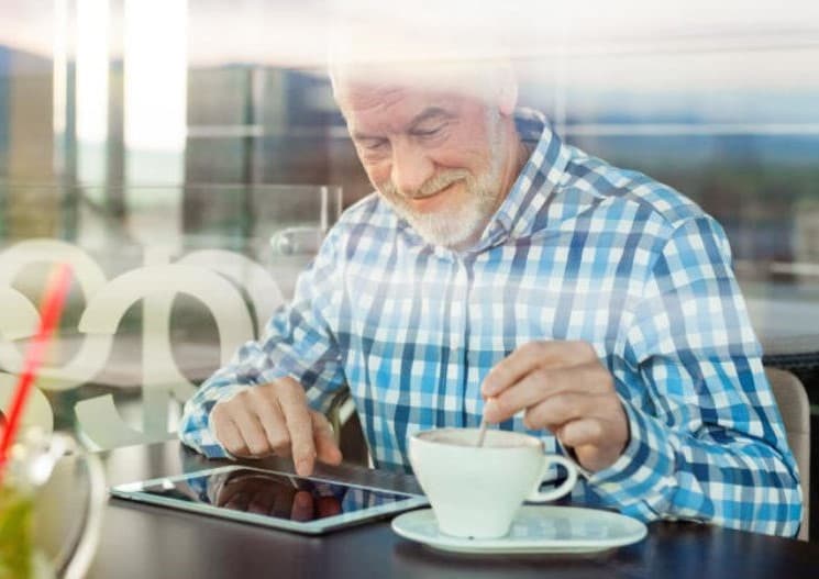 Prostatakrebspatient sitzt in Café und liest einen Newsletter auf dem Tablet während er seinen Kaffee umrührt