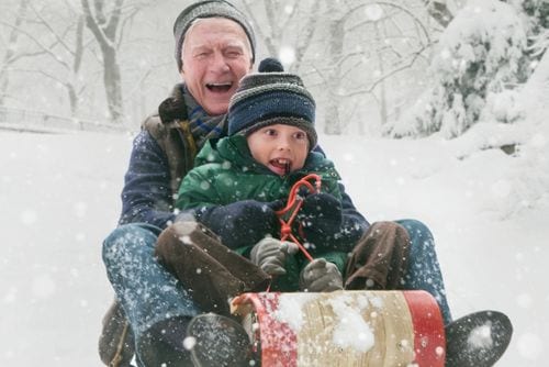 wertvollER: Prostatakrebspatient fährt lachend mit seinem Enkel auf einem Schlitten in der verschneiten Winterlandschaft