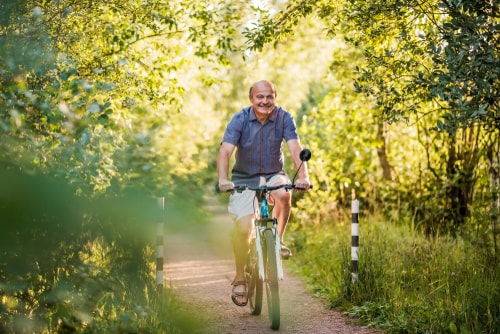 Älterer Mann fährt entspannt Fahrrad auf einem schmalen, von grünen Bäumen und Sträuchern gesäumten Weg bei sonnigem Wetter.