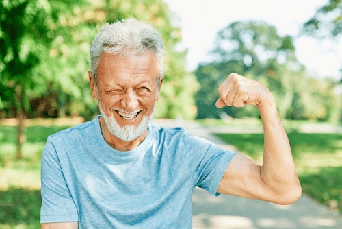 Glücklicher Senior mit grauen Haaren zeigt seinen Muskel im Park und strahlt Vitalität, Gesundheit und Fitness aus.