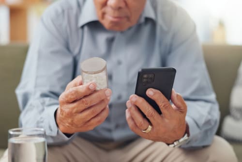 Älterer Mann hält in der linken Hand sein Smartphone und in der rechten Hand eine Medikamentendose